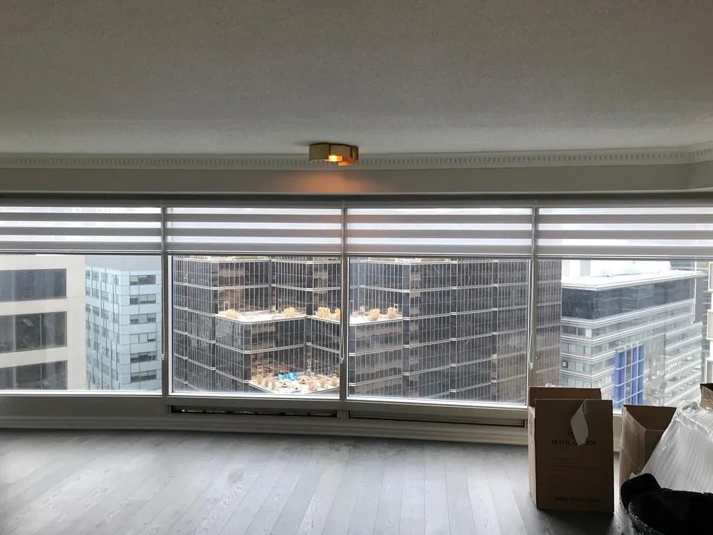 Zebra shades rolled part way up across the floor-to-ceiling glass of a high-rise flat, towers outside, moving boxes on the floor